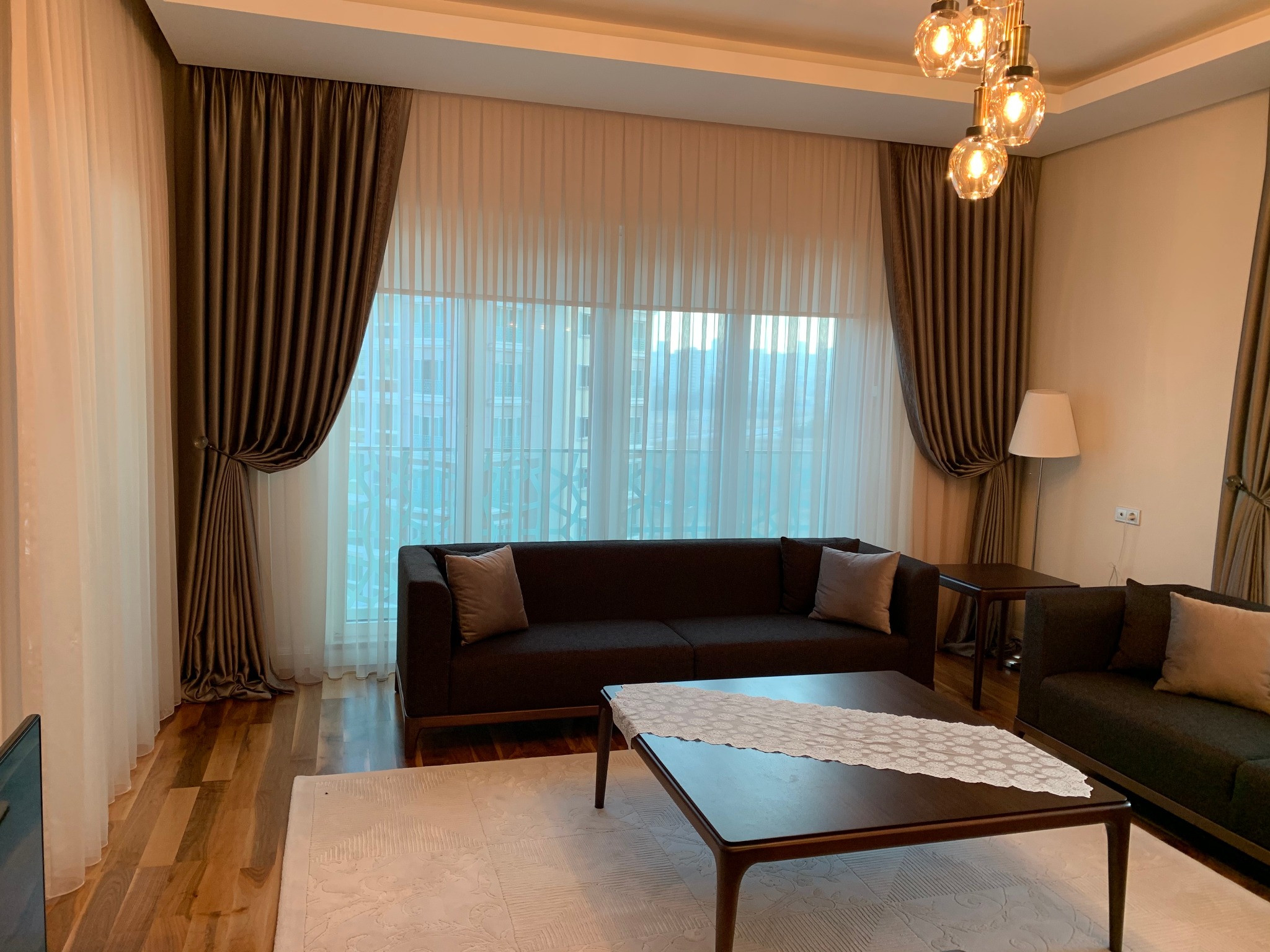 Sofa Loca salonunun oturma bölümünde geniş cam boyunca dökümlü tül ve iki yana toplanmış fon perdeler
